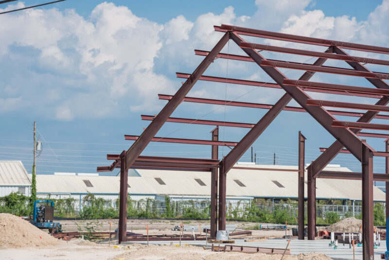 Steel structure of new industrial building under clear blue sky. New technology structural frame beam of factory in construction. Steel frame manufacturer and pile of sand and gravel in Crosby, TX, US